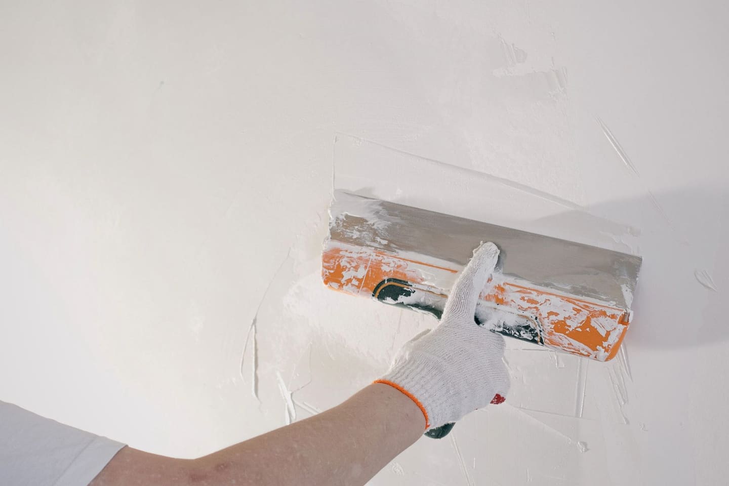 Main gantée passant un couteau à enduire sur un mur blanc.