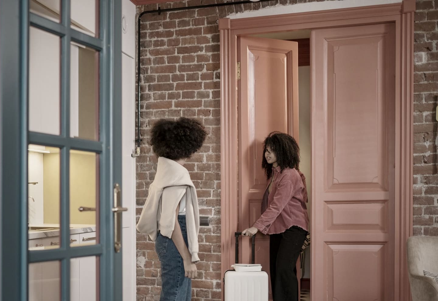 Deux voyageuses entrent dans un appartement avec une valise à roulettes, porte ouverte sur un mur de brique.
